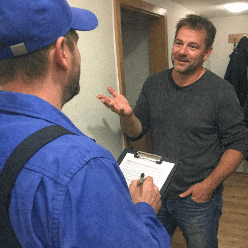 Kunde und Techniker stehen an einer Werkbank, daneben liegt eine aufgeschlüsselte Leistungsübersicht mit Positionen und Gesamtbetrag